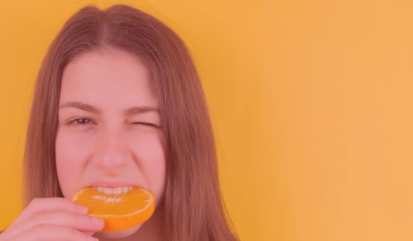 Junge Frau mit langen Haaren beißt zwinkernd in eine halbierte Orange, vor orangefarbenem Hintergrund