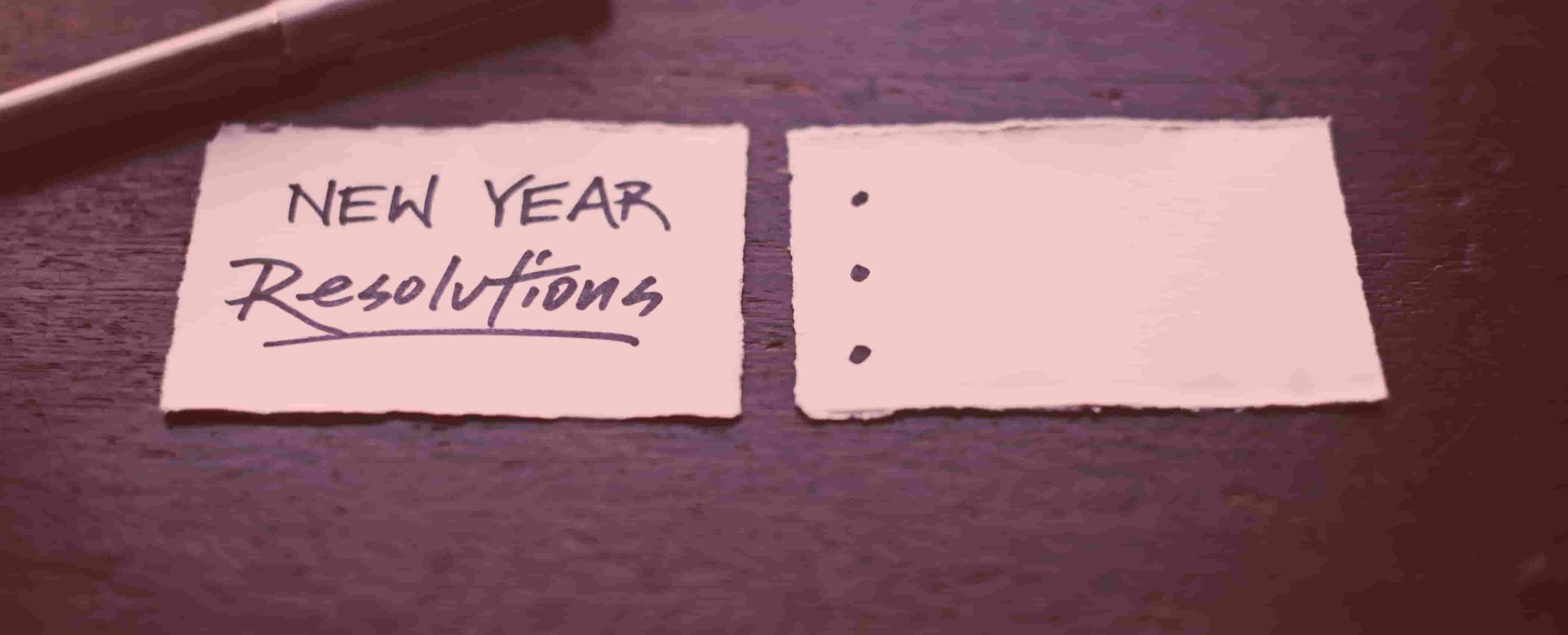 Zwei Zettel auf einem Holztisch: links steht handschriftlich „New Year Resolutions“, rechts stehen drei leere Aufzählungspunkte
