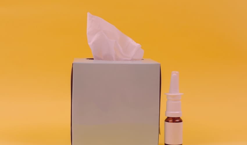 Taschentuchbox mit herausgezogenem Tuch neben einem Nasenspray, vor gelbem Hintergrund